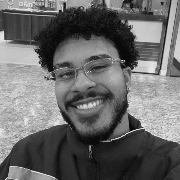 Foto em preto e branco de Matheus, jovem negro de cabelo cacheado, curto e escuro. Ele tem barba e bigode, usa óculos, veste uma jaqueta e sorri para a foto