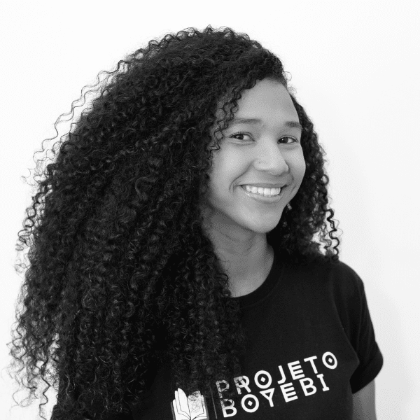 Foto em preto e branco de Hagata, jovem negra de cabelos cacheados, escuros e volumosos. Ela veste uma camiseta preta do Projeto Boyebi e sorri para a foto