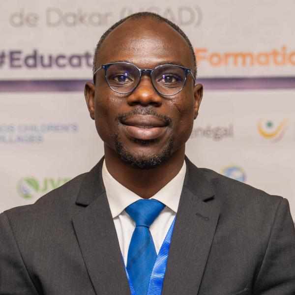 Headshot for Tchanlandjou Kpare Ashoka Sahel Staff. Person with dark brown skin and glasses smiling at the camera. Dressed in a black suit and blue tie; background of an event poster