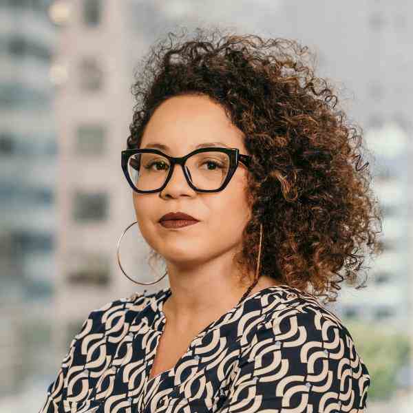 Photo of Aline, a black woman with curly hair, large hoop earrings, and dark-rimmed glasses, posing in front of a softly blurred cityscape. She wears a navy and white geometric patterned blouse and has a calm, self-assured expression with neutral makeup and burgundy lipstick.