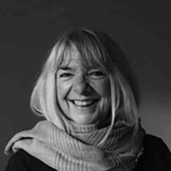 Black and white headshot of Ann Mortifee, ASN member in Canada. Person with light shoulder length hair smiling at the camera dressed in a scarf and jacket. Background is a light hued wall