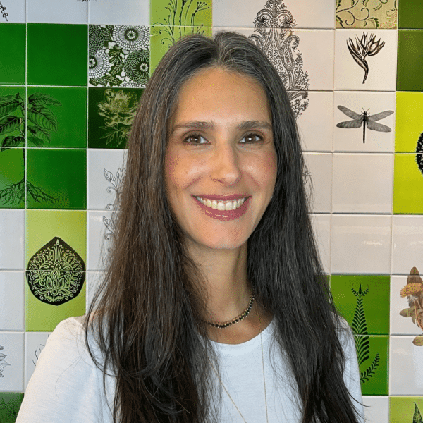 A photo of Maria Fernanda with long, straight, dark hair is pictured from the shoulders up, standing in front of a tiled wall with green and white tiles featuring botanical and insect illustrations. She is wearing a light-colored top and a simple necklace, looking directly at the camera with a smile of her face.