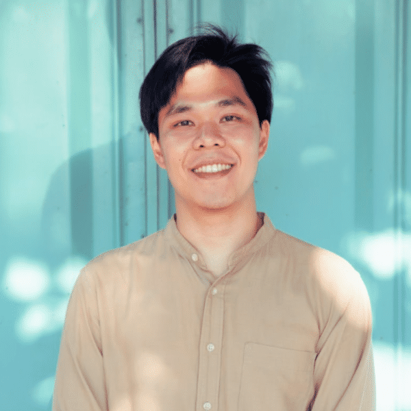 Person with short dark hair and a beige collared shirt smiling at the camera. Background a teal wall outdoors