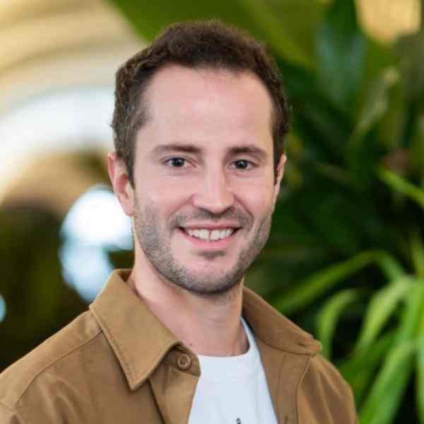 Romain Gustot staff for Ashoka Belgium. Person with short dark hair and lighter skin smiling at the camera. Dressed in a brown blazer with white shirt underneath backdrop of plants