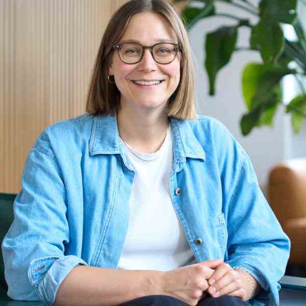 Person with shoulder length hair and glasses smiling at the camera. Seated in an office dressed in light blue collared shirt with white blouse and dark pants