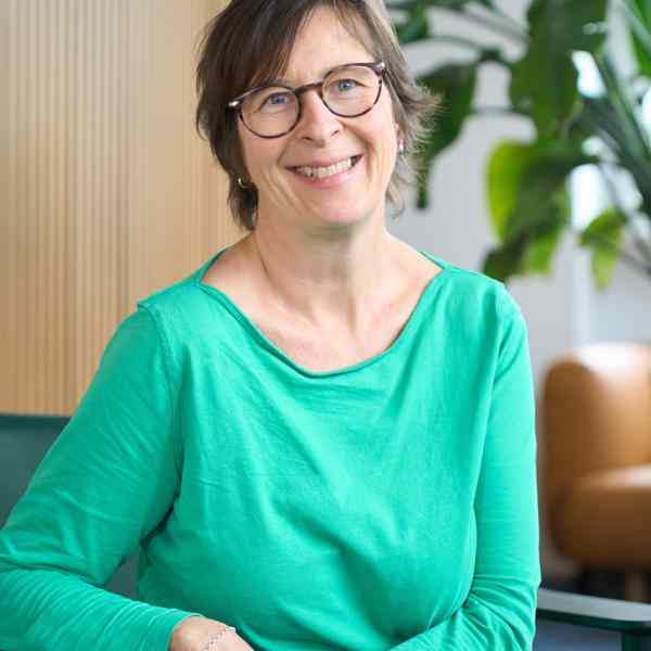 Photo of person with short length dark hair and glasses smiling at the camera. Dressed in light green shirt; background an office space