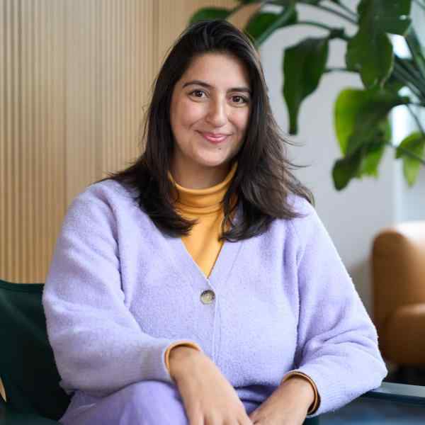 Person with shoulder length dark hair smiling at the camera. Dressed in purple blouse and pants, background is an office space