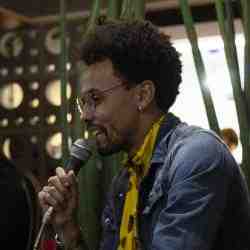 Homem negro, com cabelo black power, cavanhaque, vestindo camisa amarela e jaqueta jeans, segurando um microfone e falando.
