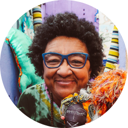 Mulher de óculos azuis, cabelo crespo, com leve sorriso, segurando uma boneca colorida.