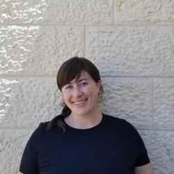Photo of Rachel Fauber, Ashoka Communications Staff. person with dark hair smiling at the camera with a black shirt on. Background is a wall made of Jerusalem stone