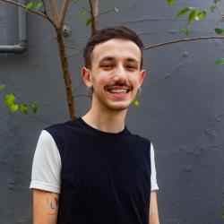 White man smiling, with a brown medium hair and a small mustache. He's wearing a black t-shirt with white sleeves. The background is a gray wall with a thin plant trunk.