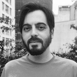 Black and white picture of Flavio Bassi. He is wearing a T-shirt, has a full beard and short dark hair.