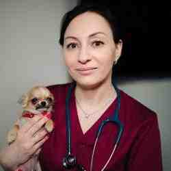 Portret Natalii Perevalovej w bordowym kitlu medycznym. Na szyi ma zawieszony stetoskop. W prawej ręcej trzyma małego brązowego pieska. Natalia ma brązowe, związane włosy, uśmiecha się.