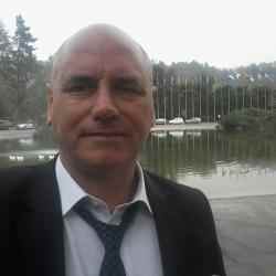 White man, wearing black suit. There is a lake in the background and some trees