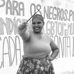 Beatriz is a black girl with shaved hair. She is wearing a T-shirt and a flowery skirt and she is holding a brush. Behind her there is a mural painted by her. The photo is black and white