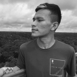 Luiz Henrique é um jovem indígena. Ele está vestindo uma camiseta e olhando para o horizonte. Ao fundo está o céu e a Floresta Amazônica