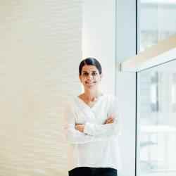 Photo of Ana Saenz de Miera; person dressed in a white shirt smiling at the camera, arms crossed. Background of a corner in between a white wall and a large window overlooking a street