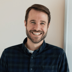 Headshot of Nick Boedicker. Person with lighter skin short hair and beard smiling at the camera. Dressed in a dark shirt, background of a wall