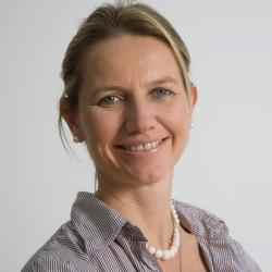 Headshot of Ashoka Fellow in Austria Katharina Kruppa. Person with lighter skin and light brown dark blonde hair tied up with a hair tie smiling at the camera. Dressed in light grey jacket. Background of a white wall