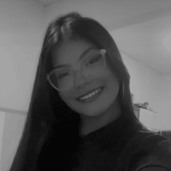 Foto em preto e branco de Ana Karoline, jovem de cabelos longos e lisos. Ela usa óculos e sorri para a foto