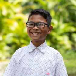 Headshot of AYC Bhre grinning cheerfully wearing a white polkadot shirt on a nature background.