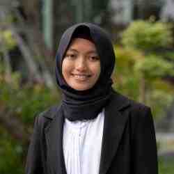 Headshot of AYC Diah smiling directly at the camera wearing a black hijab and a black suit on a nature background.