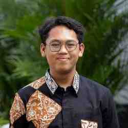 Headshot of AYC Fahry smiling directly at the camera wearing a black batik shirt on a nature background.