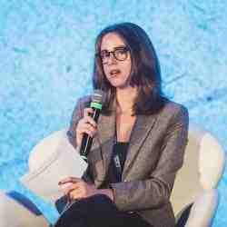 Laura Batalla of the Ashoka Belgium team. Person with dark brown shoulder length hair and lighter skin speaking into a microphone while seated in a padded chair. Dressed in a blazer and black blouse; background of light electric blue wall; chair is white