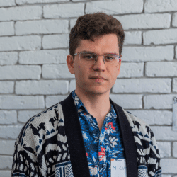 Young man in colorfull shirt and a jumper, waring glasses, standing in front of a brick wall