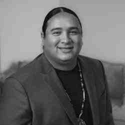 The image is a black and white photograph of Nick Tilsen wearing a dark blazer over a light-colored shirt. He has his hair tied back, so you can only see the top of his hair. Nick is wearing a necklace with a large pendant. He is smiling in the photo.