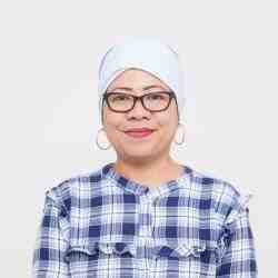 Person with white head covering and blue flannel shirt and jeans smiling at the camera. Seated on a white chair with a white background