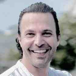 Profile photo of Ashoka Framework Change Staff Edward Edilbi. Photo of person with long slick backed hair smiling at the camera; background is of blurred out trees