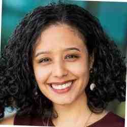Ana Saldarriaga, COO of We Are All Human Foundation, smiles warmly at the camera. She has curly black hair, wears a sleeveless maroon top and round earrings, with a softly blurred blue and green background.
