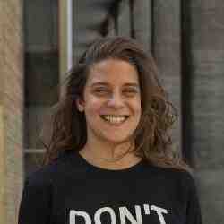 A photo of Maria Vitória: a woman with long, dark blonde, and wavy hair. She is standing outdoors. Her eyes are blue and she is smiling. Maria Vitória is wearing a black shirt with the partially visible text 'DON'T' in white letters. The background includes a building with vertical lines and windows.