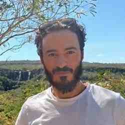 Yuri, a white man with short black hair, a beard, and a mustache, is standing outdoors wearing a white t-shirt and sunglasses on his head. In the background, there is a scenic waterfall surrounded by lush greenery under a clear blue sky.