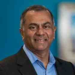 Portrait of a smiling man wearing a blue shirt and dark blazer, standing in front of a blue background.