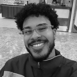 Foto em preto e branco de Matheus, jovem negro de cabelo cacheado, curto e escuro. Ele tem barba e bigode, usa óculos, veste uma jaqueta e sorri para a foto
