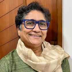 Headshot of Dr. Sujata Khandekar. She is an Indian woman with short curly hair wearing purple glasses and a tan scarf.