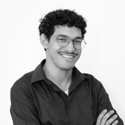 Foto em preto e branco de Nicholas, um jovem negro de pele clara com cabelo curto, escuro e cacheado. Ele tem bigode, usa óculos e veste uma camisa social cinza escuro. Nicholas está de braços cruzados e sorri para a foto