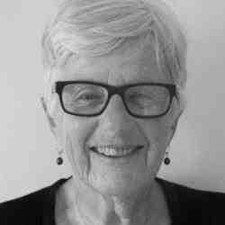 Black and white headshot off ASN Canada member Sue Griggs. Person with glasses and shorter gray hair smiling at the camera. Background a light hued wall