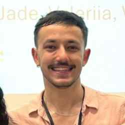 Head-and-shoulders portrait of Ricardo, a person with short dark hair, a mustache, and stud earrings, wearing a light peach shirt and a necklace, standing in front of a light background with partially visible text.