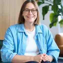 Person with shoulder length hair and glasses smiling at the camera. Seated in an office dressed in light blue collared shirt with white blouse and dark pants