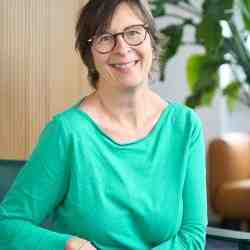 Photo of person with short length dark hair and glasses smiling at the camera. Dressed in light green shirt; background an office space