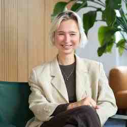 Person with short ear length blonde hair smiling at the camera. Dressed in a white blazer and dark pants. Background an office space