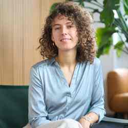 Person with shoulder length brown curly hair smiling at the camera. Dressed in blue dress shirt and white pants. Background an office space