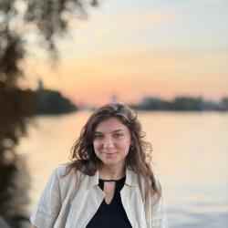 Person with shoulder length light brown hair smiling at the camera; dressed in black blouse with white collared shirt over it. Background a sunset over a river