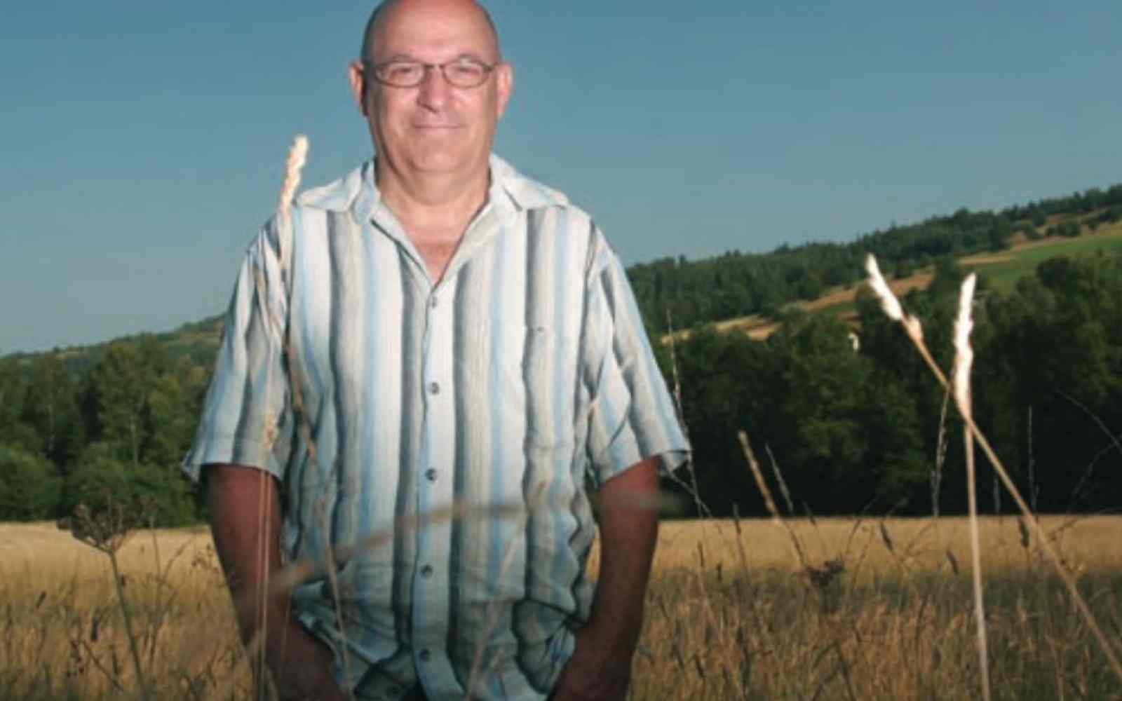 Zdjęcia Piotra Topińskiego na tle pola. Piotr ubrany jest w białą koszulę w pionowe pasy. Uśmiecha się.