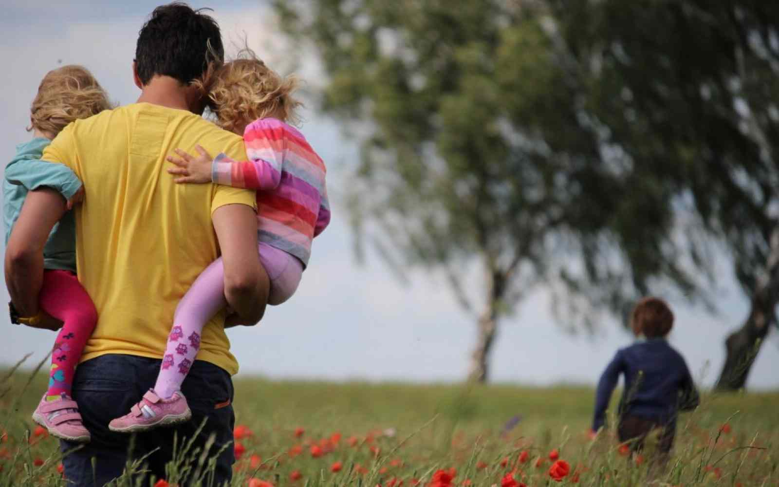 Zdjęcie pokazuje od tyłu tatę w żółtej koszulce trzymającego na rękach dwoje dzieci. Przed nimi biegnie jeszcze jedno, starsze dziecko. Wspólnie idą przez łąkę. W tle duże drzewo.