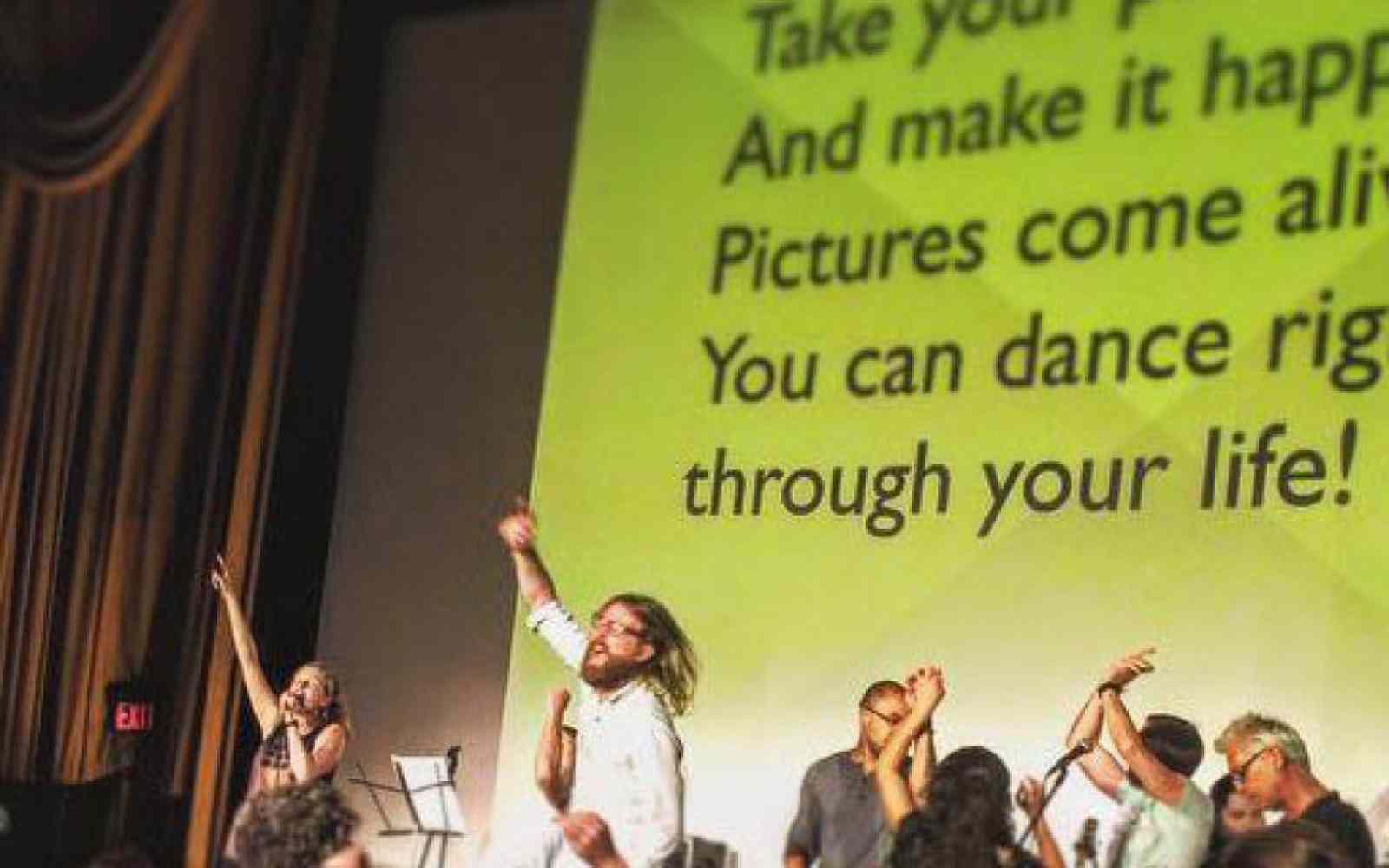 Zdjęcie przedstawia występ zespołu na scenie podczas Sunday Assembly. W tle na ekranie rzutnika wyświetlany jest tekst piosenki.
