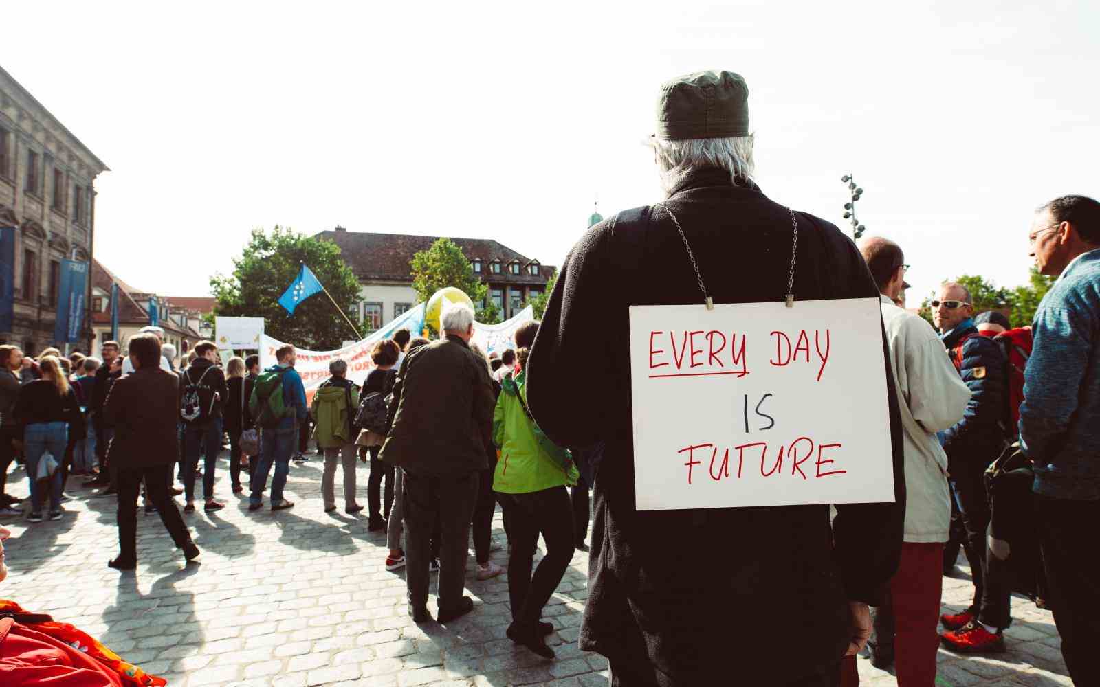 Mężczyzna w ciemnej kurtce ma na plecach zawieszony baner z napisem "every day is future". W tle widać innych protestujących,